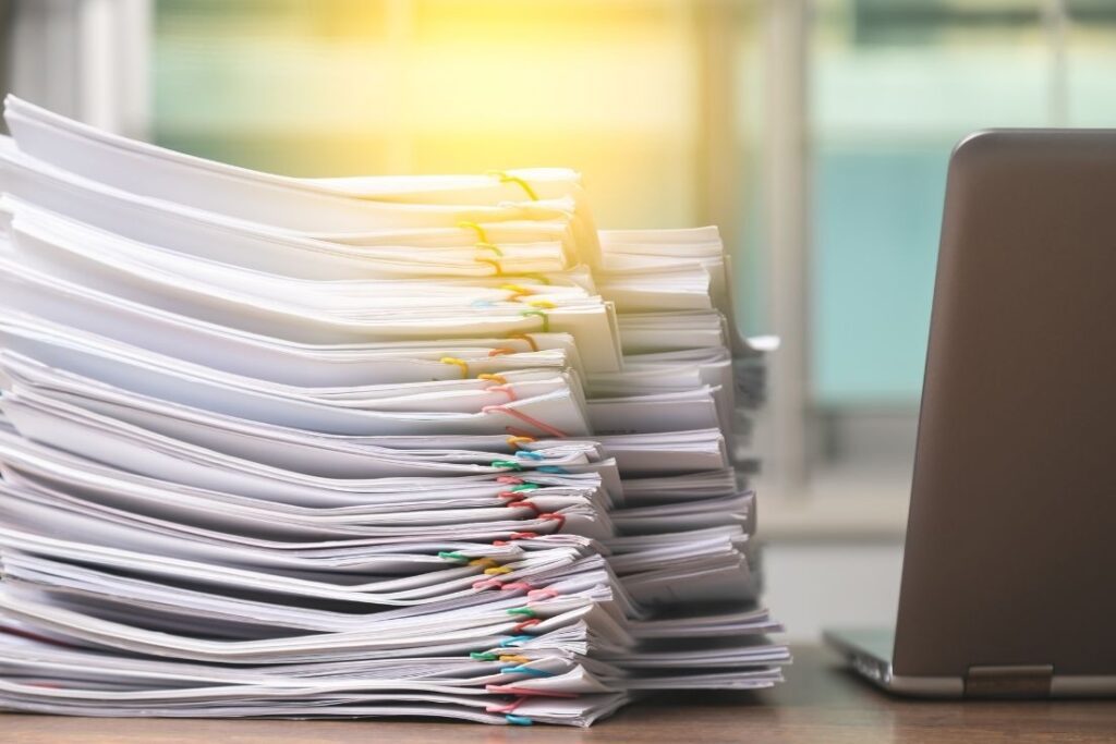 Pile of paper documents stacked beside a laptop, representing a cluttered office environment with paper-based workflows.