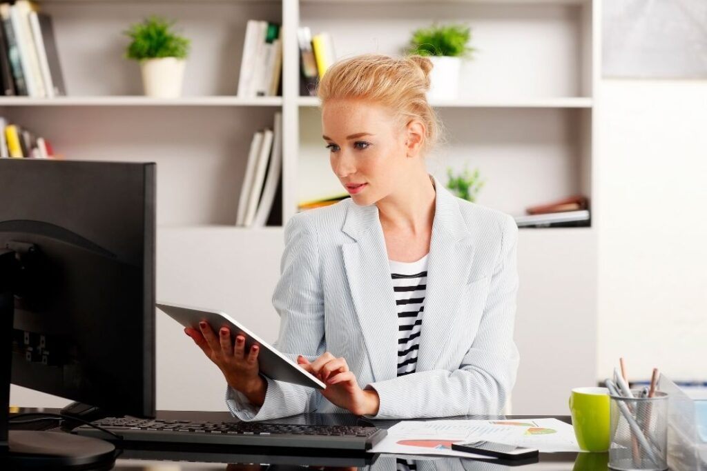 Professional woman using a tablet while sitting at a desk, with a computer monitor and organizational items in the background. She appears focused and engaged in work.