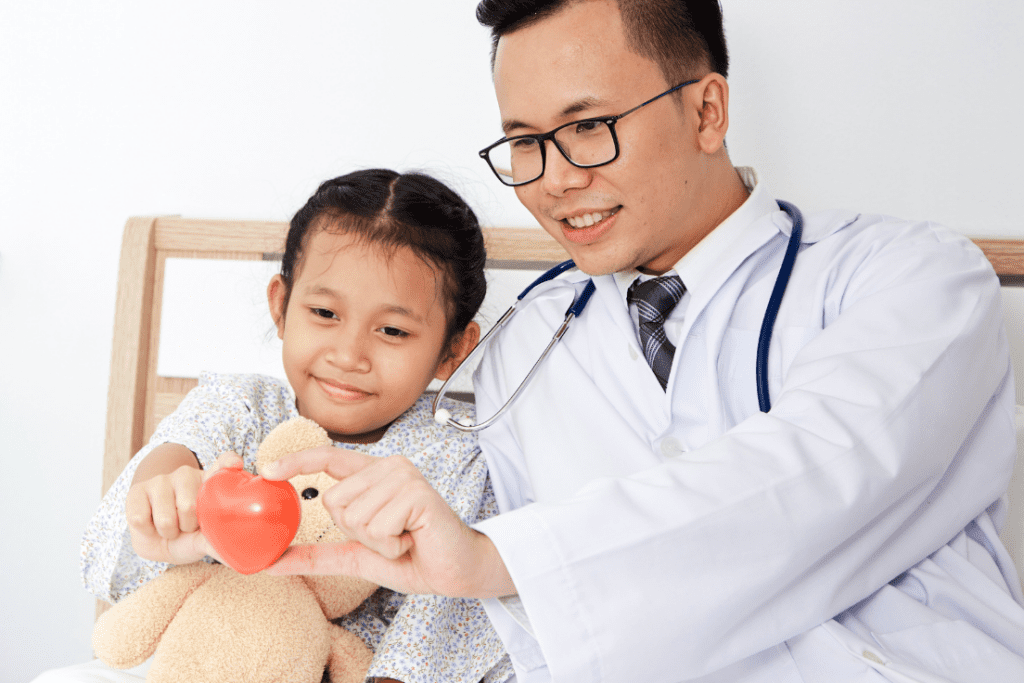 Pediatric doctor and young girl holding a heart-shaped object, smiling and interacting together. The doctor is wearing a stethoscope, and the girl is holding a teddy bear.