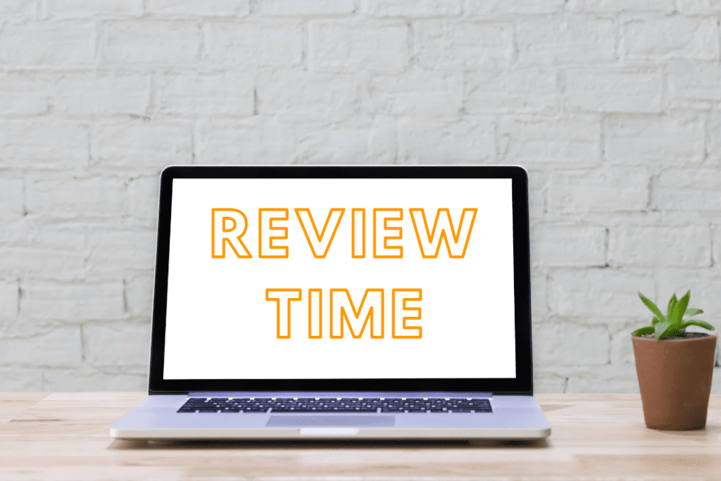 A laptop on a desk displaying the text "REVIEW TIME" on its screen, with a small potted plant next to it and a white brick wall in the background.