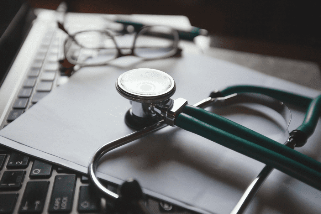 A stethoscope, glasses, and paperwork on top of a laptop keyboard, symbolizing the combination of healthcare management and administrative tasks.