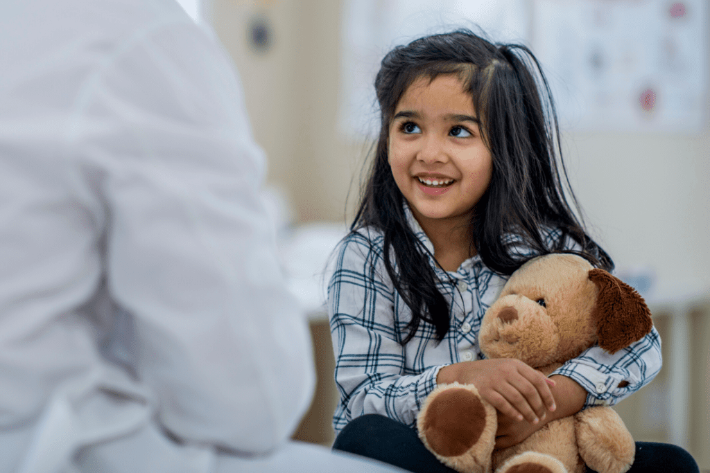 A young girl holding a teddy bear, smiling at a healthcare professional, representing patient care and comfort in healthcare settings.