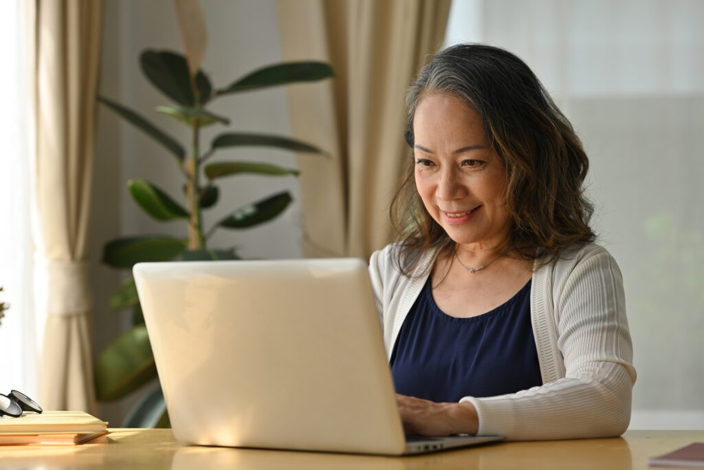 An elderly woman is smiling while working on her laptop at a table in a well-lit room with plants.