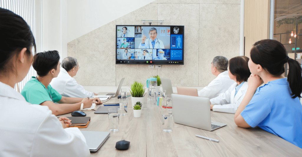 A group of healthcare professionals attending a virtual committee meeting via video conference. They are sitting around a table with laptops, tablets, and notebooks, engaging with a doctor on the screen.