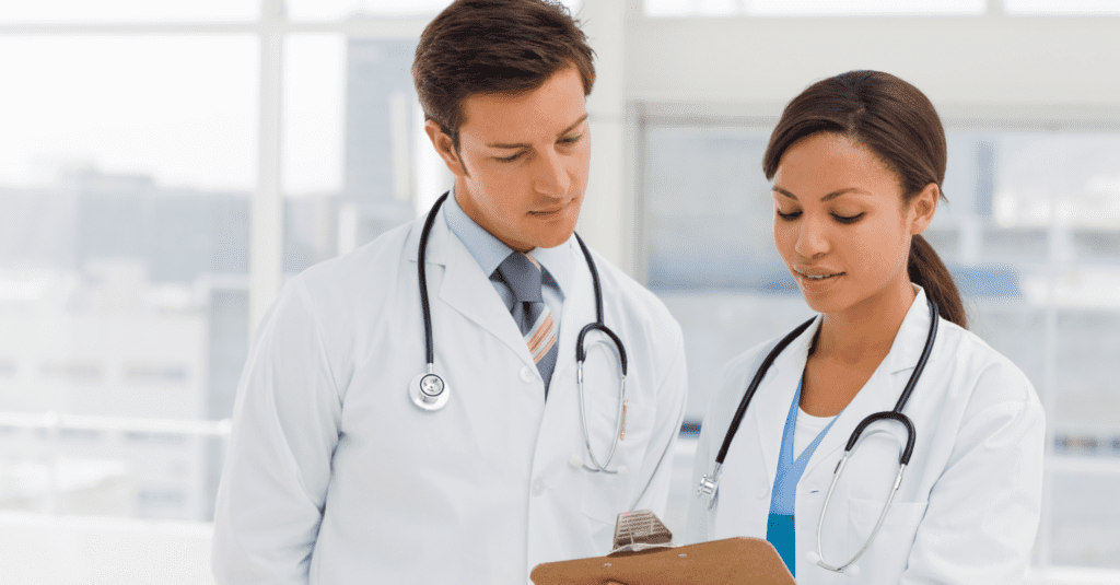 A male and female doctor are reviewing medical documents on a clipboard in a bright hospital or office setting.