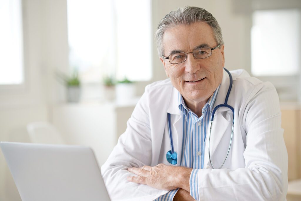 An elderly doctor wearing glasses and a white coat, smiling and sitting in front of a laptop in a bright office.