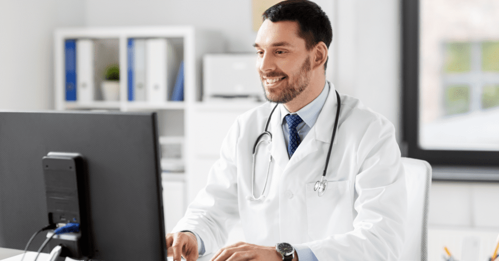 A smiling male doctor in a white coat working on a computer in a medical office.