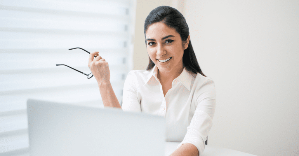 A woman sitting at a desk with a laptop, smiling while holding her glasses, symbolizing ease and professionalism.