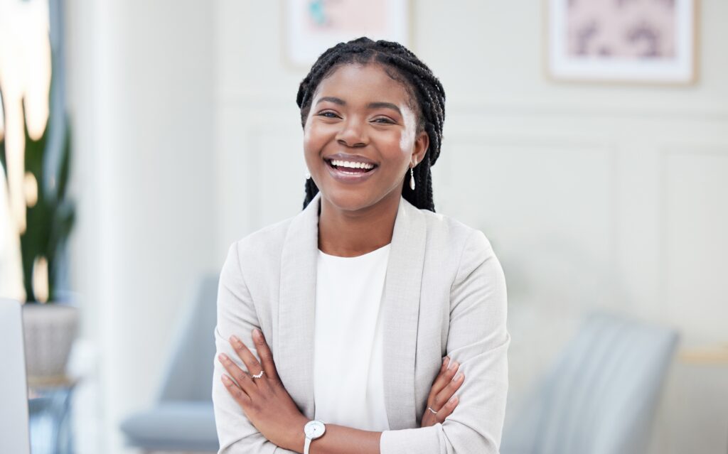 A young woman with braided hair, smiling and standing with arms crossed in a modern office environment.