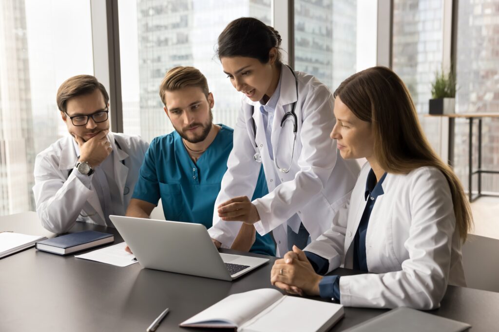 Group of doctors collaborating in a modern office, with one pointing at a laptop screen while others observe and engage in a discussion.