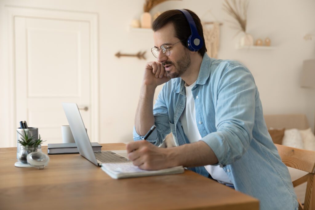 Man studying at home, wearing headphones and glasses, taking notes while working on his laptop.