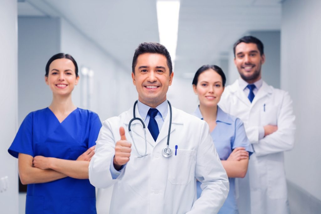 A diverse group of healthcare professionals standing together in a hospital corridor, smiling and giving a thumbs up.