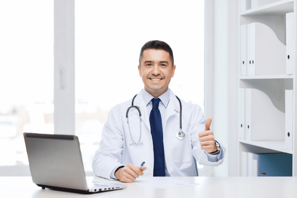 Smiling doctor giving a thumbs-up while sitting at a desk with a laptop in a bright office.