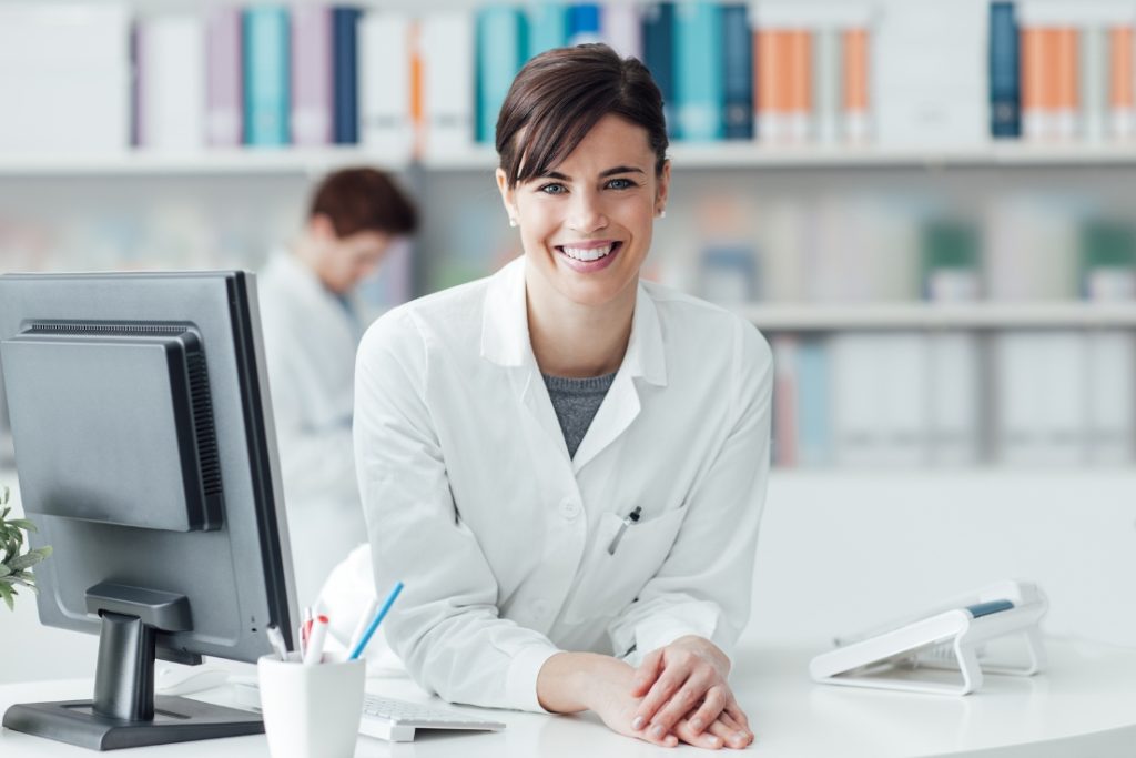A smiling female healthcare professional in a white coat at her desk with a computer, representing efficient credentialing processes.