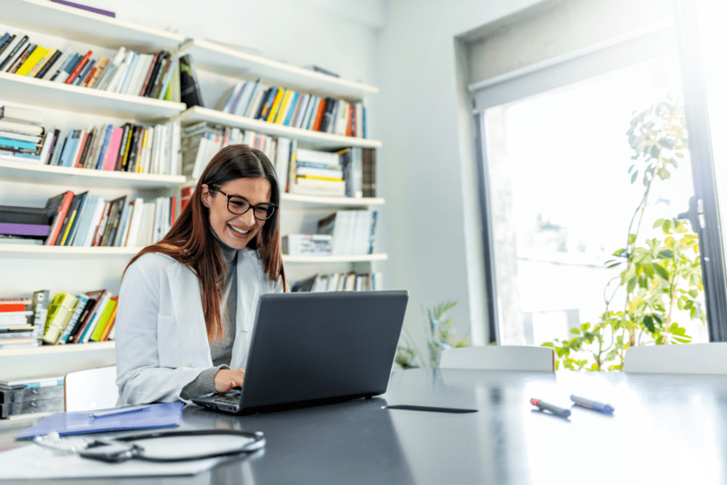 A healthcare professional in a white coat smiling while working on a laptop in a modern office, with bookshelves and medical documents in the background.