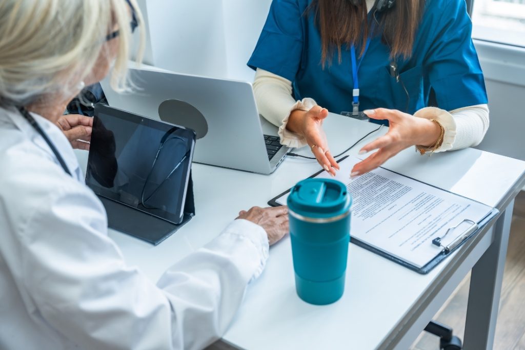 Two people in front of their computers discussing paperwork.