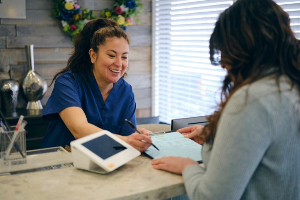 Image of two women in a medical office smiling and handing over paperwork.